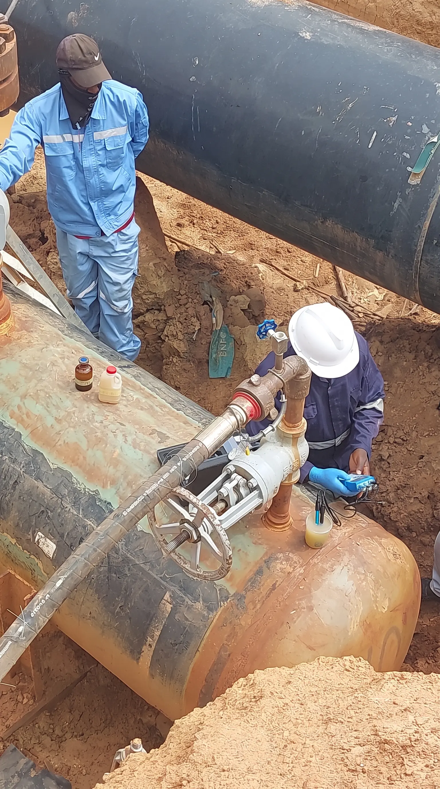 Technicians using testing equipment on an exposed pipeline in an excavated trench.