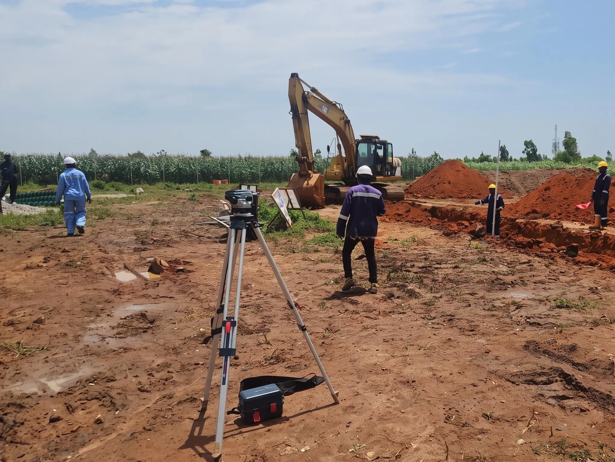 Land surveying team using a tripod-mounted level and measuring staff beside an excavated trench and excavator.